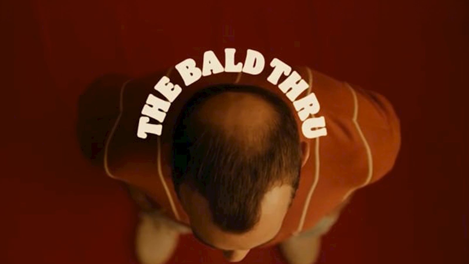 A birds eye view of a balding man standing on a red floor with the text "The Bald Thru" in white