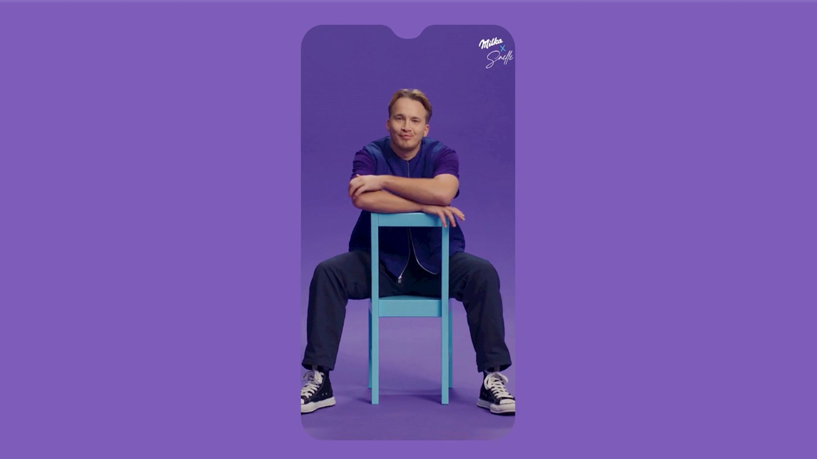 Purple background with a phone screen imposed on to it with Dutch musician Snelle sitting backwards on a light blue chair