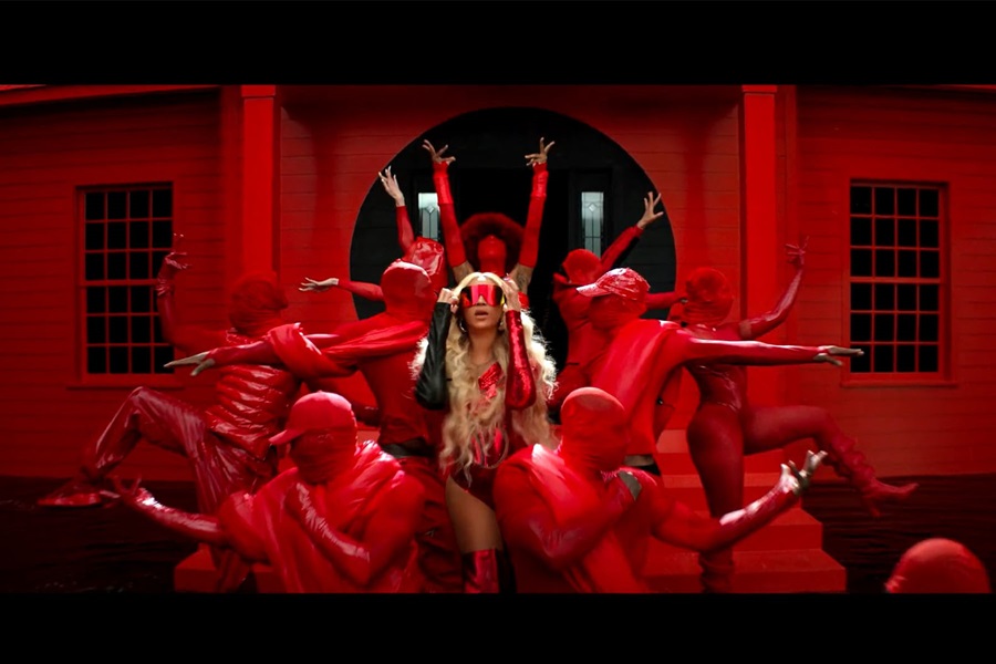 Beyonce dressed in red putting red sunglasses on her face, standing in front of a red house amidst a group on dancers also dressed in red