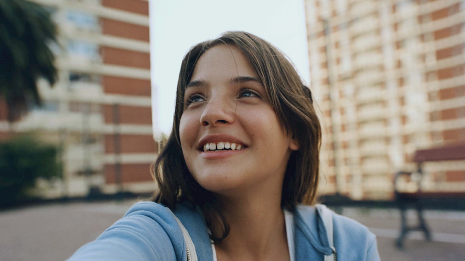 Young girl with brown hair wearing a blue hoodie, sitting in front of an apartment block, looking up to the right and smiling