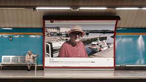 Elderly woman sitting in a subway station looking at a large billboard with a photo of herself on it